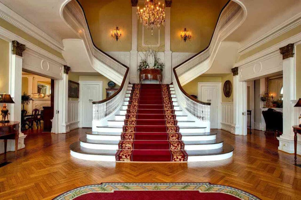Modern interior design_img (1) Elegant grand foyer with a split staircase adorned with a red carpet and intricate gold detailing. A sparkling chandelier enhances the luxurious ambiance.
