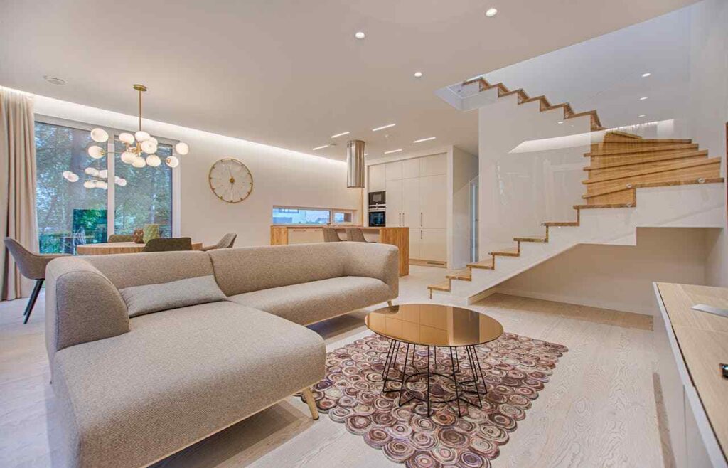Modern living room with a beige sofa, round coffee table, and abstract rug. Elegant staircase and dining area with pendant lights in the background.
