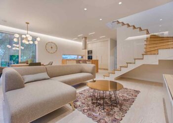 Modern living room with a beige sofa, round coffee table, and abstract rug. Elegant staircase and dining area with pendant lights in the background.