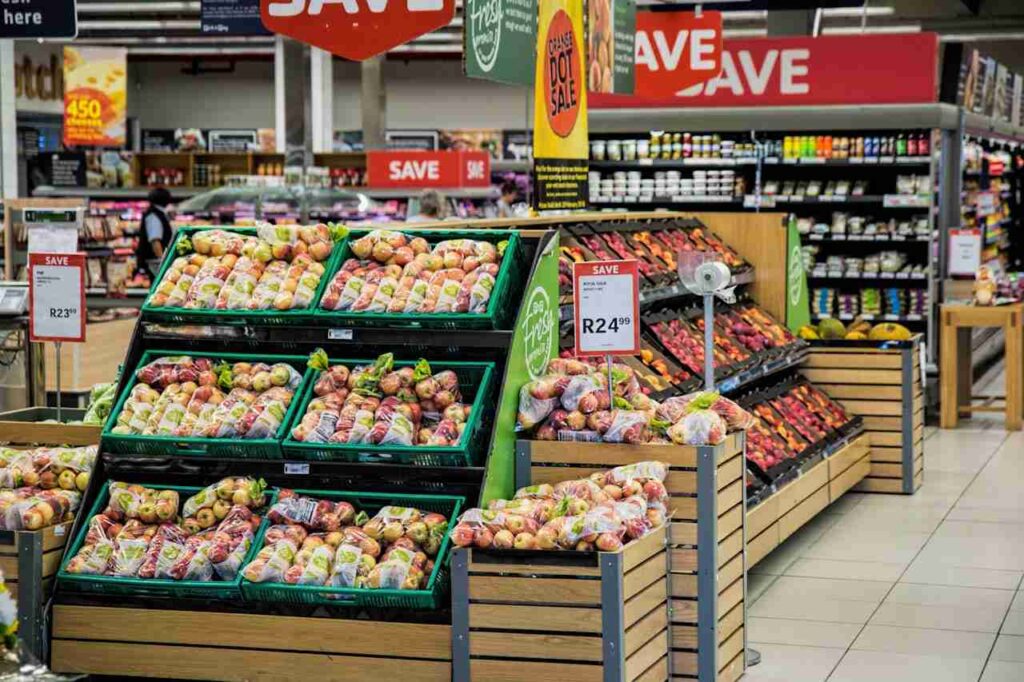 a grocery store with 6 boxes of fruits on 3 shelves, a red sign that says save, other vegetables on shelves in the background, and a fridge containing drinks.