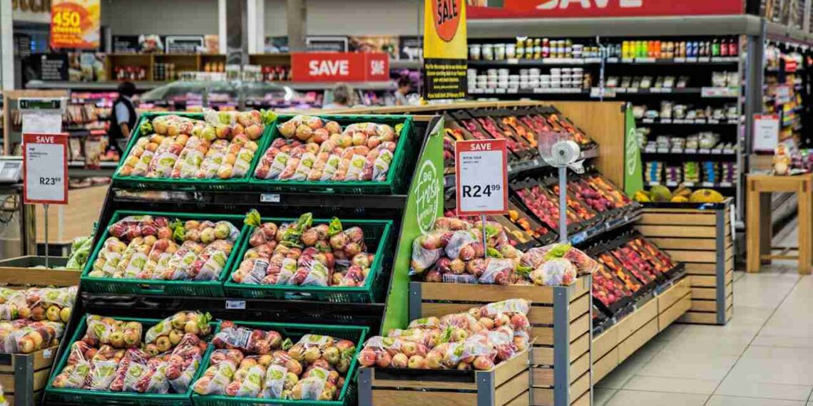 a grocery store with 6 boxes of fruits on 3 shelves, a red sign that says save, other vegetables on shelves in the background, and a fridge containing drinks.