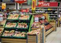 a grocery store with 6 boxes of fruits on 3 shelves, a red sign that says save, other vegetables on shelves in the background, and a fridge containing drinks.