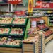 a grocery store with 6 boxes of fruits on 3 shelves, a red sign that says save, other vegetables on shelves in the background, and a fridge containing drinks.